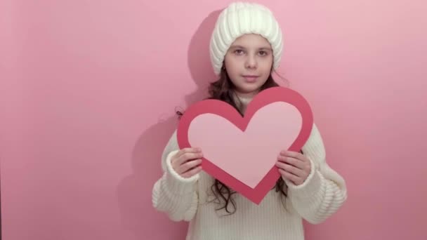 Jeune fille souriante avec un coeur en papier fait à la main dans ses mains sur fond rose — Video