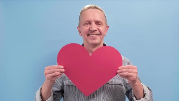 Lächelnd Alter Mann zeigen großes rotes Herz isoliert blaue Farbe Hintergrund. — Stockvideo