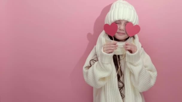 Happy Young girl wearing white knitted sweater holding red hearts over eyes — Stock Video
