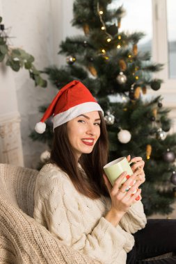 Gülümseyen genç bir kızın elinde yeşil bir fincan kakao tutarken Noel şenliği ağacının yanında.