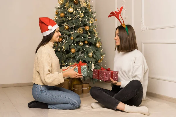 Dvě krásné dívky v bílých svetrech dát si navzájem červené a zelené vánoční dárky — Stock fotografie