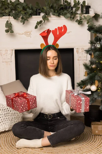 Молода дівчина з пухкими губами сидить і тримає два червоних різдвяних подарунки біля білого каміна Ліцензійні Стокові Зображення
