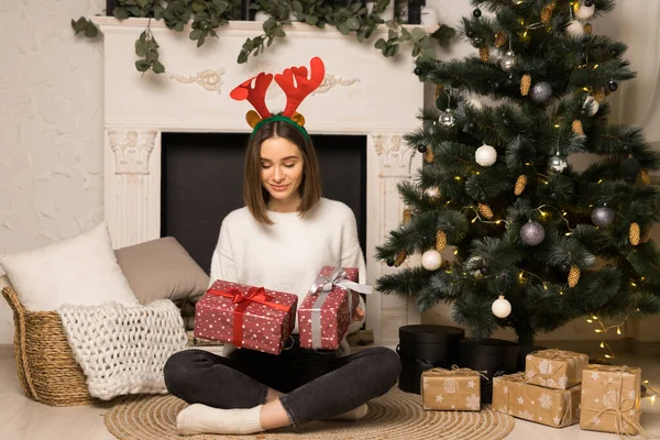 Здивована молода дівчина сидить і дивиться на два червоних різдвяних подарунки біля білого каміна Стокова Картинка