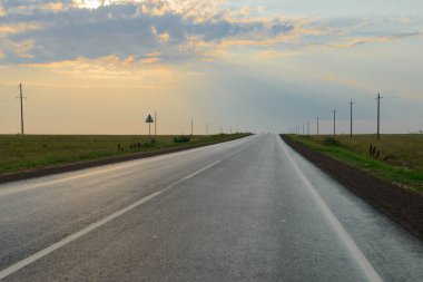 Arazinin ortasında asfalt bir yol. Güneş ışınları bulutların arasından geçiyor.