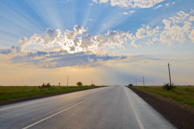 Arazinin ortasında asfalt bir yol. Güneş ışınları bulutların arasından geçiyor.