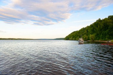 Geniş sakin bir nehri, sahilde bir ormanı ve eski bir teknesi olan bir yaz manzarası.