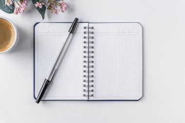 Beyaz yüzeyde bir fincan kahve, not defteri, kalem ve pembe çiçek.