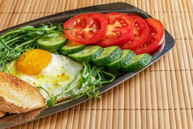 Kızarmış yumurta, kızarmış ekmek, salatalık ve domates dilimleri ve bambu saman yüzeyinde metal bir tabakta mikro yeşiller, sağlıklı kahvaltı kavramı.