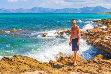 Erkek gezgin ve turist, İspanya 'nın Balearic Adası Mallorca' daki Can Picafort 'ta turkuaz su dalgaları ve kaya parçalarıyla engebeli doğal kıyı ve sahil manzarasında poz veriyor..