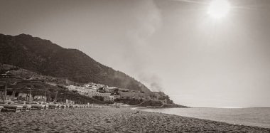 Oros Dikaios Dağları 'nın altındaki şemsiyeler, lüks tatil beldeleri ve plajların üzerinde duman ve güneşin doğuşunun eski siyah beyaz resmi Kos Adası Yunanistan mavi gökyüzü ile doğal kıyı manzaraları.