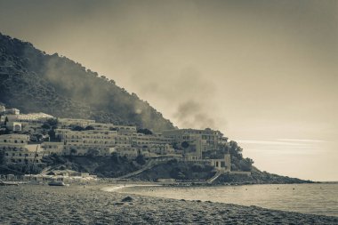 Oros Dikaios Dağları 'nın altındaki şemsiyeler, lüks tatil beldeleri ve plajların üzerinde duman ve güneşin doğuşunun eski siyah beyaz resmi Kos Adası Yunanistan mavi gökyüzü ile doğal kıyı manzaraları.