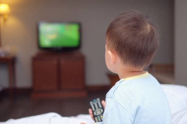 2-3 yaşlarındaki tatlı Asyalı bebek elinde uzaktan kumandayla yatakta oturan ve yatak odasında televizyon izleyen bebek, Toddlers Screen Time & Early Brain Development konsepti