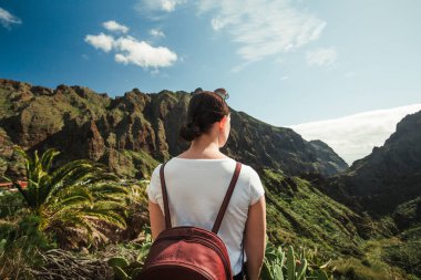 İspanya 'nın Tenerife kentindeki Masca köyüne bakan sırt çantalı bir turist. Kanarya Adaları 'nda bulutlu gökyüzü olan palmiye ağaçları ve yeşil tepelerin panoramik manzarası.