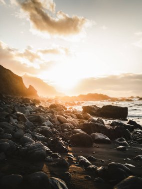 Beautiful view of rocky beach and cliff along North Tenerife coast at sunset. Atlantic wavy ocean with golden sun in Canary Islands.