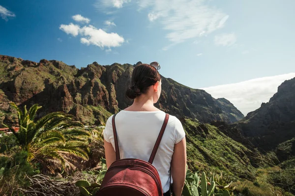 İspanya 'nın Tenerife kentindeki Masca köyüne bakan sırt çantalı bir turist. Kanarya Adaları 'nda bulutlu gökyüzü olan palmiye ağaçları ve yeşil tepelerin panoramik manzarası.