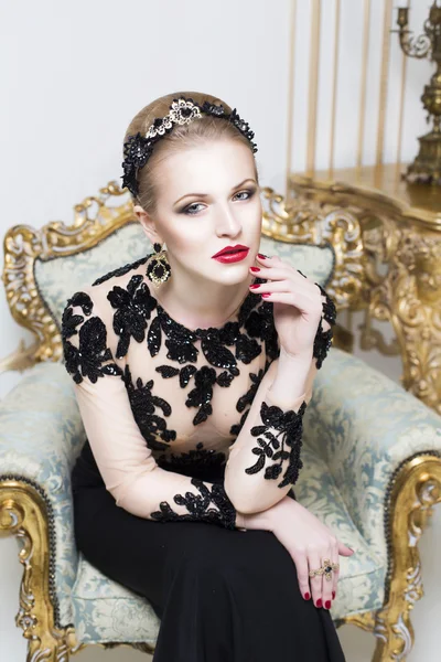 Beautiful blonde royal woman sitting on a retro chair in gorgeous luxury dress, looking at camera. Indoor — Stock Photo, Image