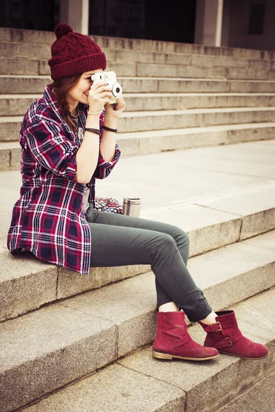 Brunette woman in hipster outfit sitting on steps on the street, photographing. Toned image