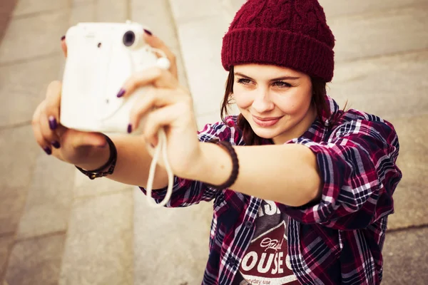 Brunette woman in hipster outfit sitting on steps and taking selfie on retro camera on the street. Toned image — Stock Photo, Image