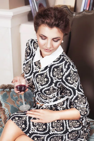 Красивая брюнетка богатая дерзкая женщина в элегантном платье сидит на стуле в комнате с классическим интерьером питьевого вина. В помещении — стоковое фото