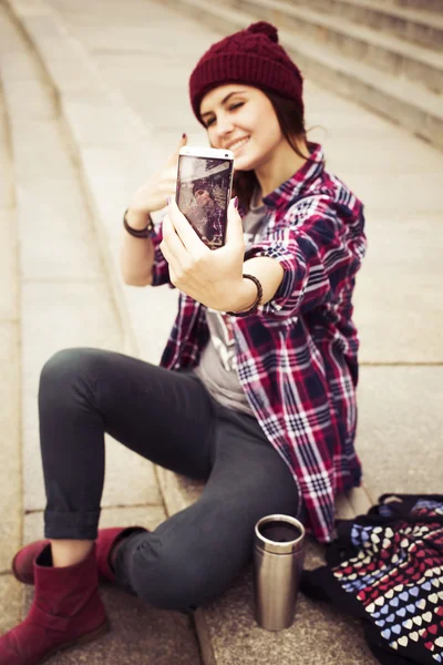 Brunette woman in hipster outfit sitting on steps and taking selfie on retro camera on the street. Toned image — Stock Photo, Image