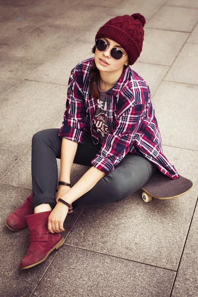 Brunette woman in hipster outfit sitting on a scateboard on the street. Toned image — Stock Photo, Image