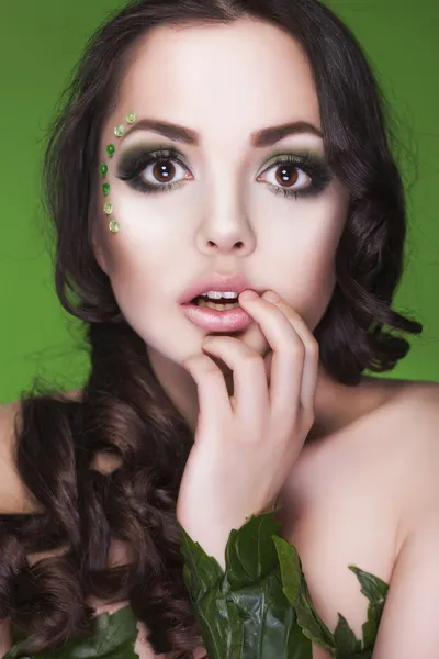 Beautiful brunette dryad woman with creative make up and beads on her face, curly hair and costume made of leaves on green background looking at camera in amazement. Hand near her lips — Stock Photo, Image
