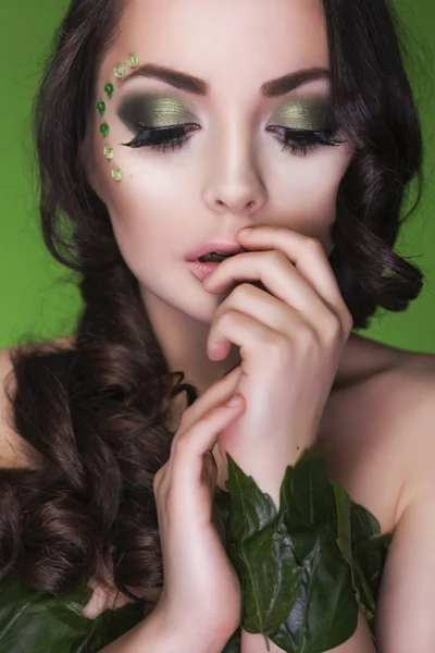 Beautiful brunette dryad woman with creative make up and beads on her face, curly hair and costume made of leaves on green background. Eyes closed — Stock Photo, Image