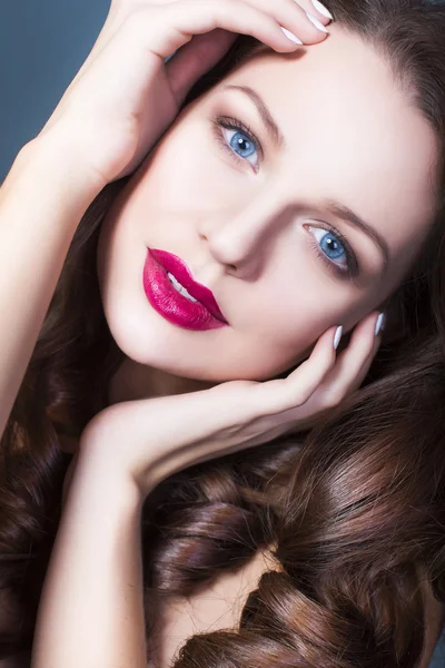 Beautiful brunette woman with creative make up violet eye shadows full red lips, blue eyes and curly hair with her hand on her face — Stock Photo, Image