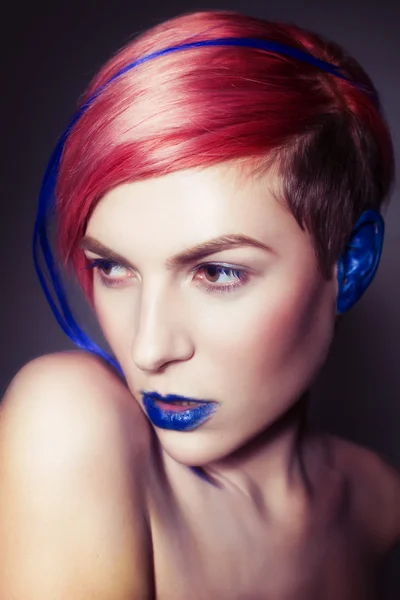 Young person with blue eye shadows, blue ears and pink hair with blue strand on it looking at camera and hands near face. Black background — Stock Photo, Image