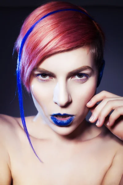 Young person with blue eye shadows, blue ears and pink hair with blue strand on it looking at camera and hands near face. Black background — Stock Photo, Image