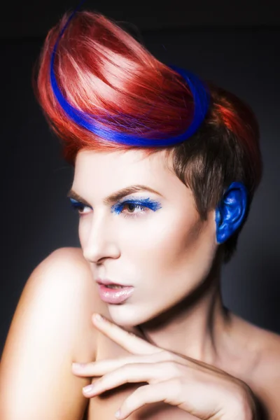 Young person with blue eye shadows, blue ears and pink hair with blue strand on it looking at camera and hands near face. Black background — Stock Photo, Image