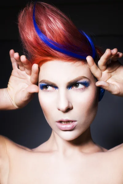 Young person with blue eye shadows, blue ears and pink hair with blue strand on it looking at camera and hands near face. Black background