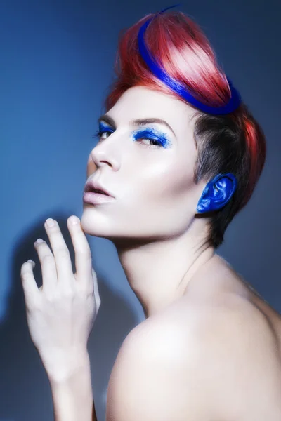 Young person with blue eye shadows, blue ears and pink hair with blue strand on it looking at camera and hands near face. Black background — Stock Photo, Image