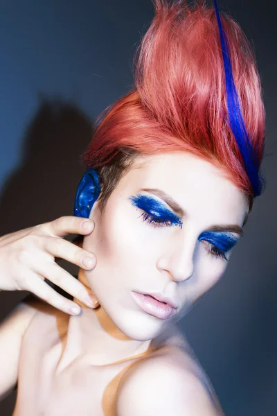 Young person with blue eye shadows, blue ears and pink hair with blue strand on it looking at camera and hands near face. Black background — Stock Photo, Image