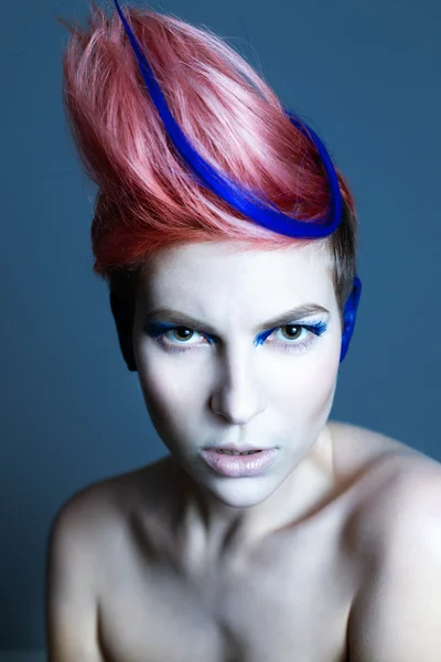 Young person with blue eye shadows, blue ears and pink hair with blue strand on it looking at camera and hands near face. Black background — Stock Photo, Image