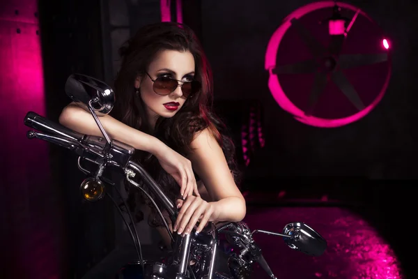 Brunette sexy woman in black underwear, heels and sunglasses in studio in red light on a motorcycle. Indoors. Copy Space — Stock Photo, Image