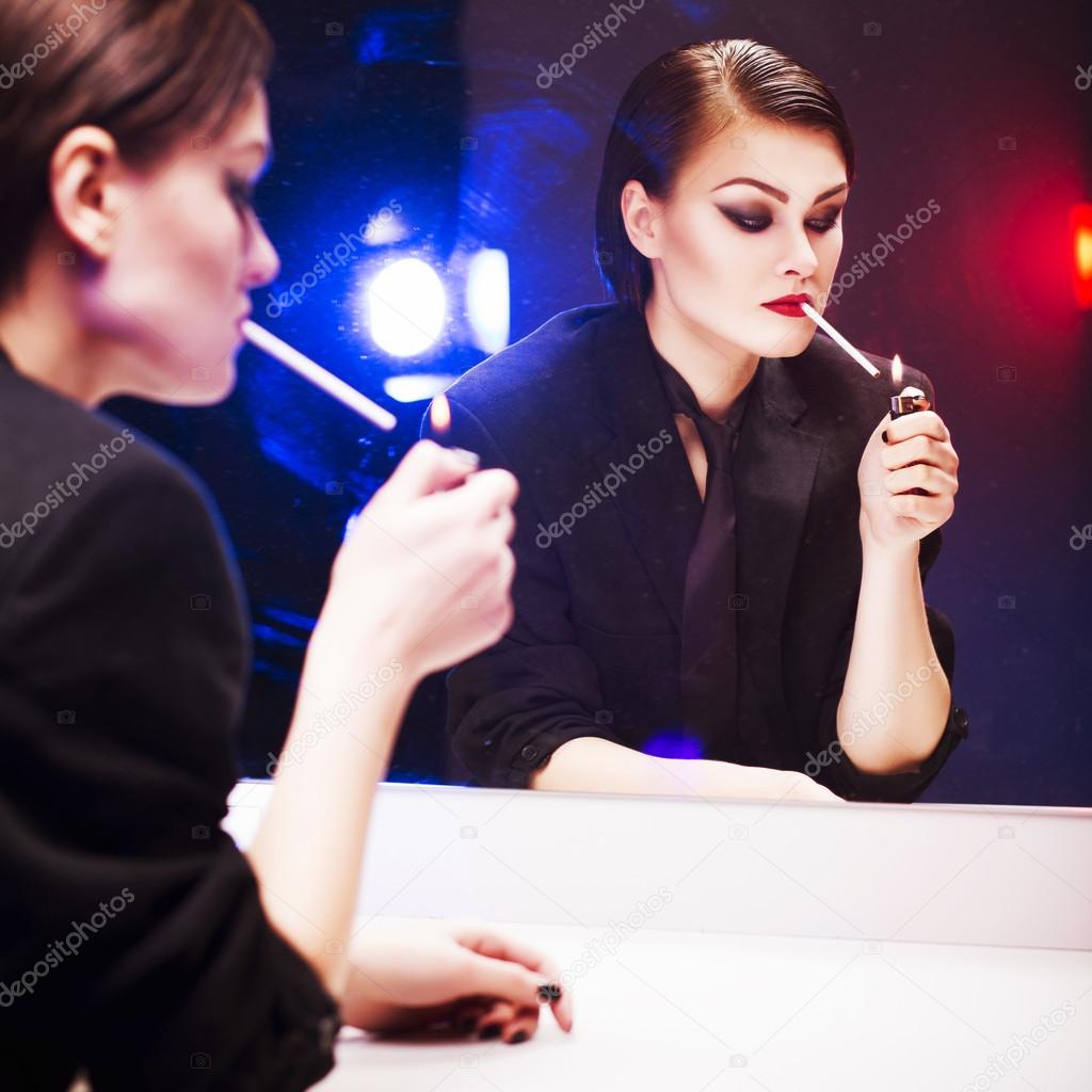 Beautiful brunette woman in business costume lighting a cigarette in front of the mirror with color backlight