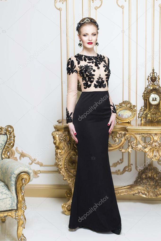 Beautiful blonde royal woman standing near retro table in gorgeous luxury dress looking in front of herself. Indoor
