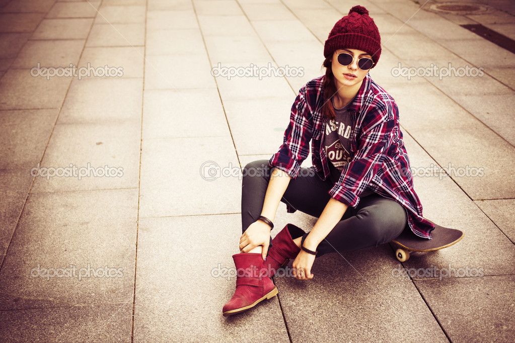 Brunette woman in hipster outfit sitting on a scateboard on the street. Toned image. copy space