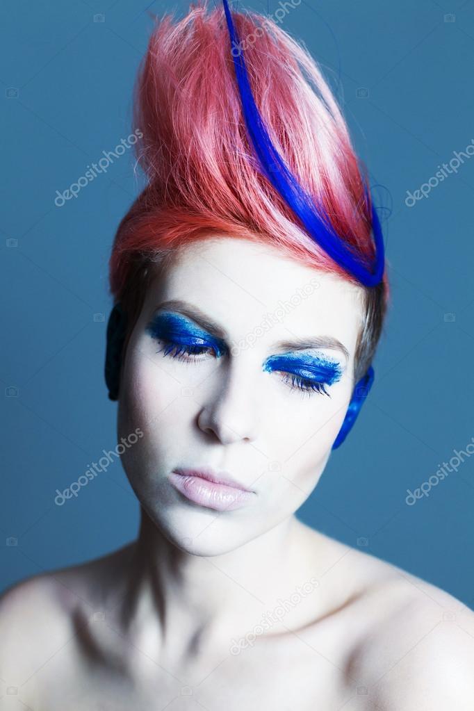 Young person with blue eye shadows, blue ears and pink hair with blue strand on it looking at camera and hands near face. Black background