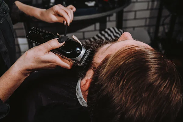 Women professional hairdresser serving client with thick big beard by clipper, handsome brutal stylish young man getting trendy haircut , black barber shop room