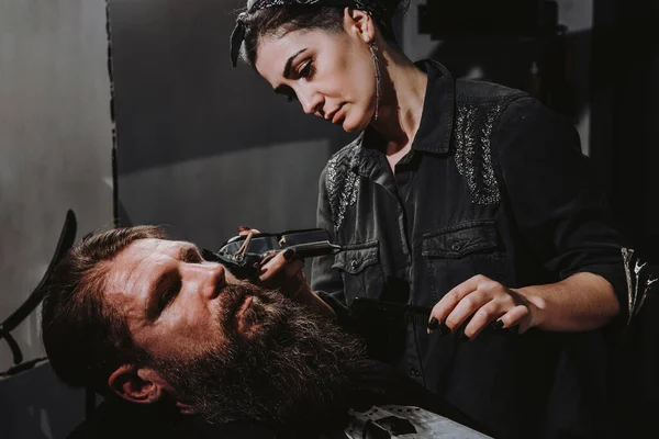 Women professional hairdresser serving client with thick big beard by clipper, handsome brutal stylish young man getting trendy haircut , black barber shop room
