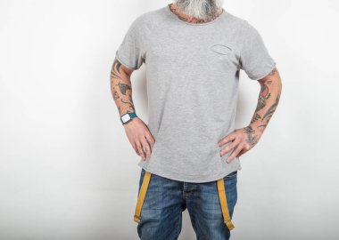 Sakallı, dövmeli bir adamın beyaz arka planda pantolon pantolon pantolon ve gri pamuklu bir tişört giyip poz verişi..