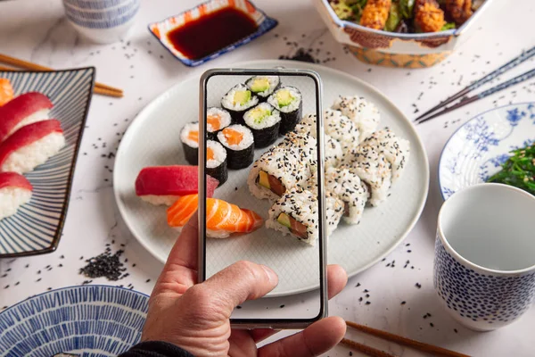 スモークサーモンと巻き巻き男性の手は携帯電話で寿司ロールの写真を撮ります スマートフォンで寿司の皿の写真を撮る若者 携帯電話での食品写真 ロイヤリティフリーのストック画像