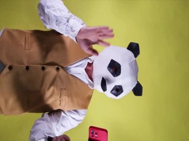 Sarı arka planda panda maskeli bir adam akıllı telefonuyla video konferansında konuşuyor. Portre biçimi videosu