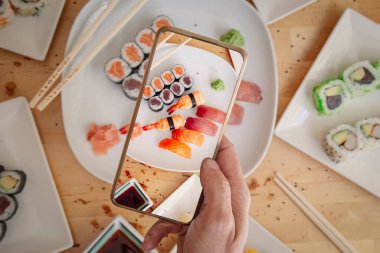 Cep telefonlu bir adam masanın üstünde lezzetli suşi fotoğrafı çekiyor.