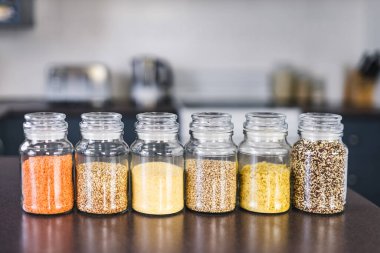 İçinde kinoa mercimek, karabuğday ve arpanın da bulunduğu farklı türde tahıl ve baklagillerin bulunduğu açık kiler kavanozları mutfak tezgahı, basit malzeme konsepti.