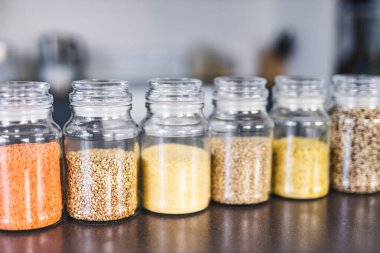 İçinde kinoa mercimek, karabuğday ve arpanın da bulunduğu farklı türde tahıl ve baklagillerin bulunduğu açık kiler kavanozları mutfak tezgahı, basit malzeme konsepti.