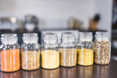İçinde kinoa mercimek, karabuğday ve arpanın da bulunduğu farklı türde tahıl ve baklagillerin bulunduğu açık kiler kavanozları mutfak tezgahı, basit malzeme konsepti.