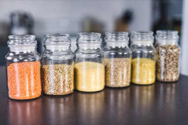 İçinde kinoa mercimek, karabuğday ve arpanın da bulunduğu farklı türde tahıl ve baklagillerin bulunduğu açık kiler kavanozları mutfak tezgahı, basit malzeme konsepti.